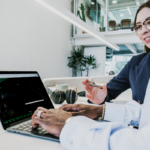 a business woman looking over a laptop with green words on a black screen, being used by a man wearing a white long sleeve shirt wearing a ring