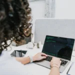 Long curly hair person typing at a laptop with green words and a black screen, over the shoulder view